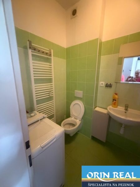 Garzonlakás fürdőszobája zöld csempével, mosdóval, WC-vel és mosógéppel.