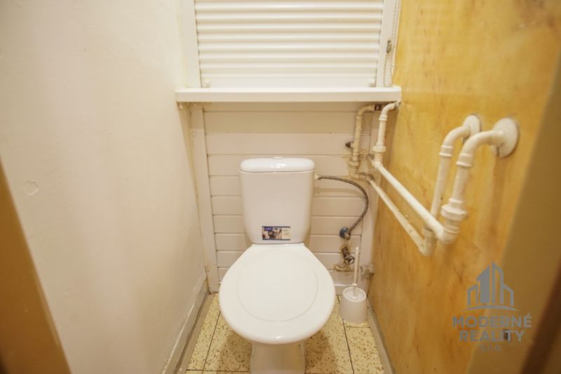 Kis fürdőszoba egy kétszobás lakásban WC-vel, fehér falakkal és oldalfalon lévő csövekkel.