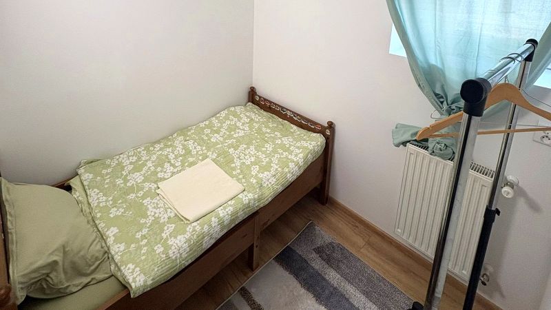 Egyszemélyes ágy, ruhatartó állvány, zöld függönyök, fa mintázatú padló.