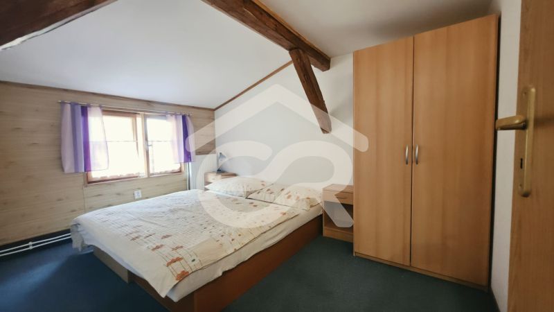 Hálószoba egy nyaraló apartmanban fa gerendákkal, szekrénnyel és ággyal az ablak mellett.