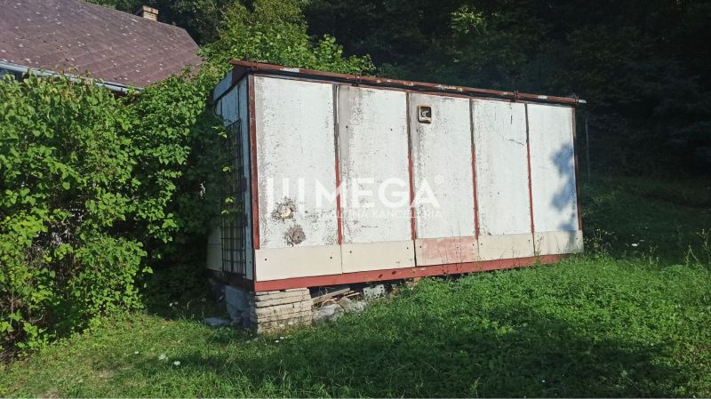 Egy régi raktár az utcai kertben Humennóban, zöldnövényzettel és növekvő fűvel körülvéve.