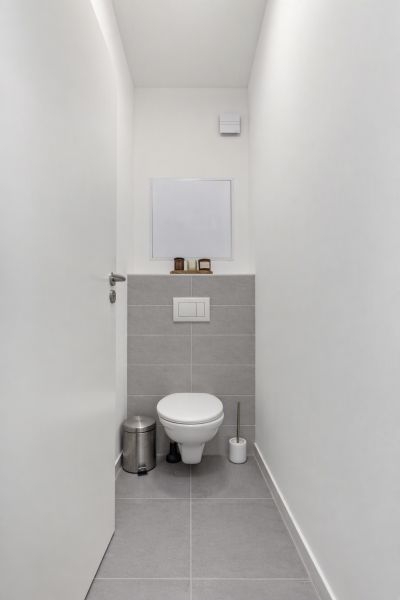 Keskeny WC fehér falakkal és szürke csempékkel egy kétszobás lakásban.