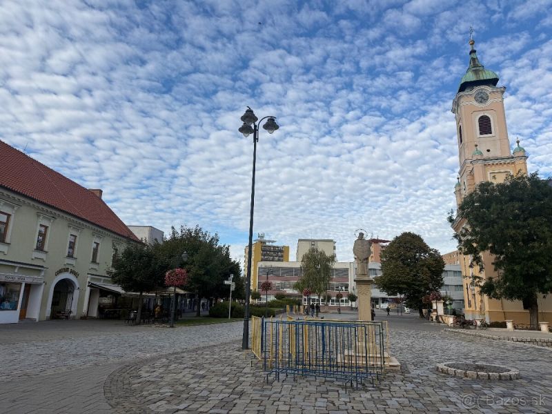 Üzlethelyiségek Topoľčanyban, kövezett térrel és háttérben egy templommal.