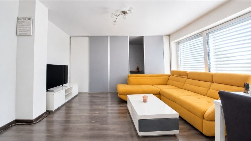 Nappali egy családi házban sárga kanapéval, TV-vel és fa mintázatú padlóval.